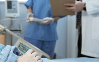 Patient in hospital bed with a nurse and doctor in the background looking at a chart.